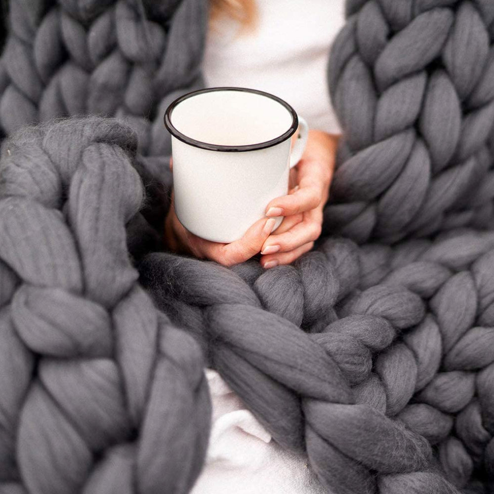 Frau hält Emaille-Tasse, eingewickelt in graue, grob gestrickte Decke aus Merinowolle.