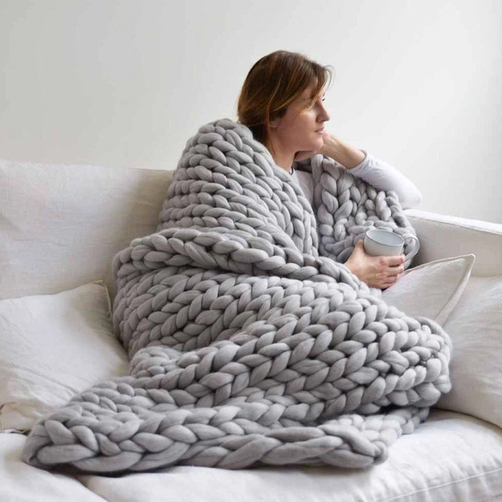 Frau mit grauer, grob gestrickter Decke auf Sofa, hält Tasse; gemütliche Wohnkultur.